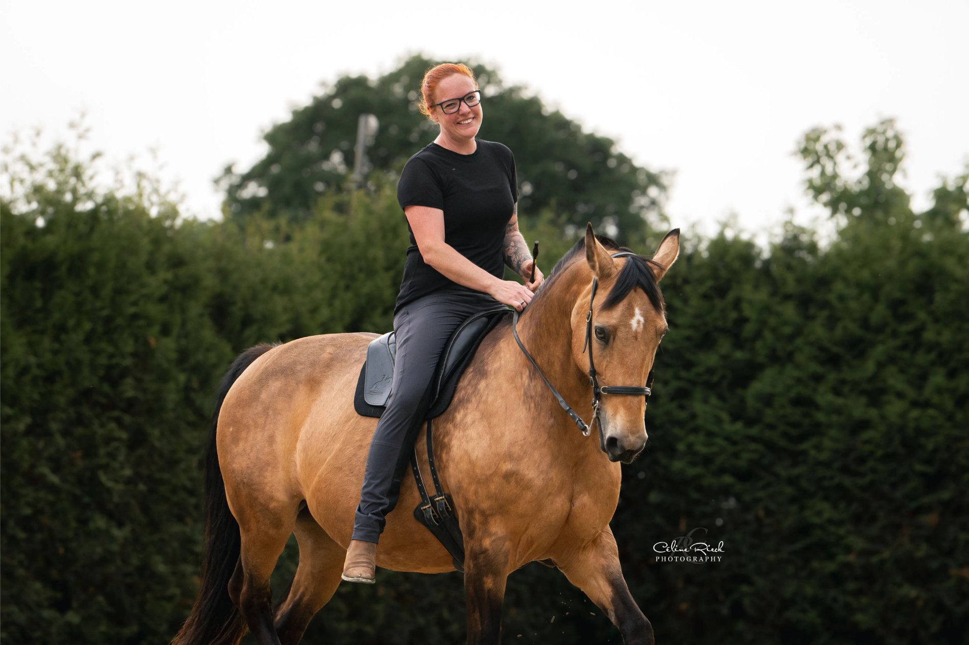 Reiterin mit roten Haaren reitet lächelnd auf einem Buckskin-Pferd im Freien – Pferdefotografie von Celine Rieck Photography