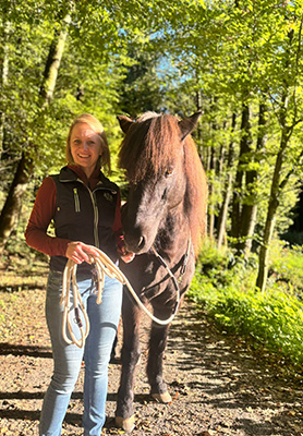 Junge Frau führt braunes Pferd am Seil durch einen sonnigen Waldweg mit grünen Bäumen im Frühling