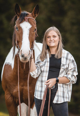Junge Frau in Karohemd posiert lächelnd mit braunem Paint Horse mit Blesse auf grüner Wiese – Pferdefotografie Outdoor