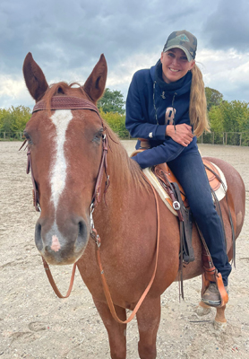 Junge Reiterin mit Kappe auf braunem Western-Pferd mit Blesse in Reitanlage bei bewölktem Himmel